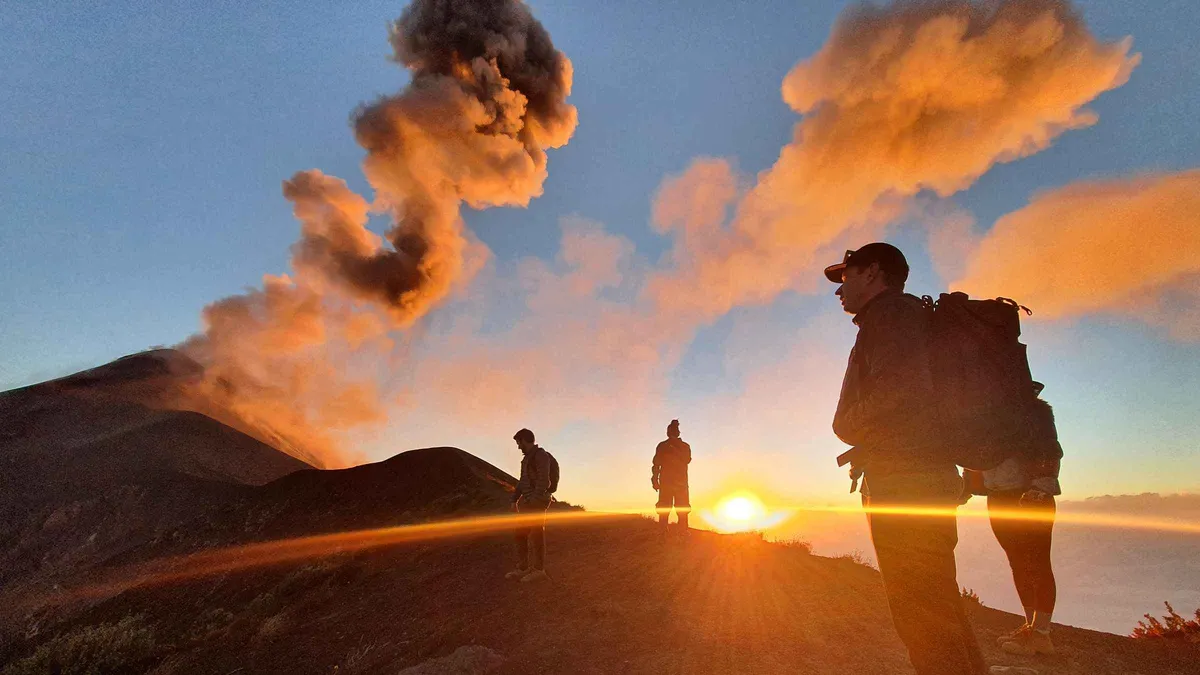Hikers silhouetted on a ridge at sunrise with an ash plume behind them on an Acatenango volcano tour above Antigua, Guatemala
